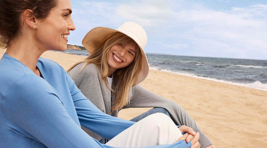Two women sit on the beach, one wears a broad brim sun hat, both wear UPF50+ sun protective clothing.